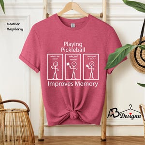 May include: Heather raspberry t-shirt with a white graphic that says "Playing Pickleball Improves Memory". The graphic features stick figures with speech bubbles that say "What's the score?", "Who just served?", and "Were you 1 or 2?"