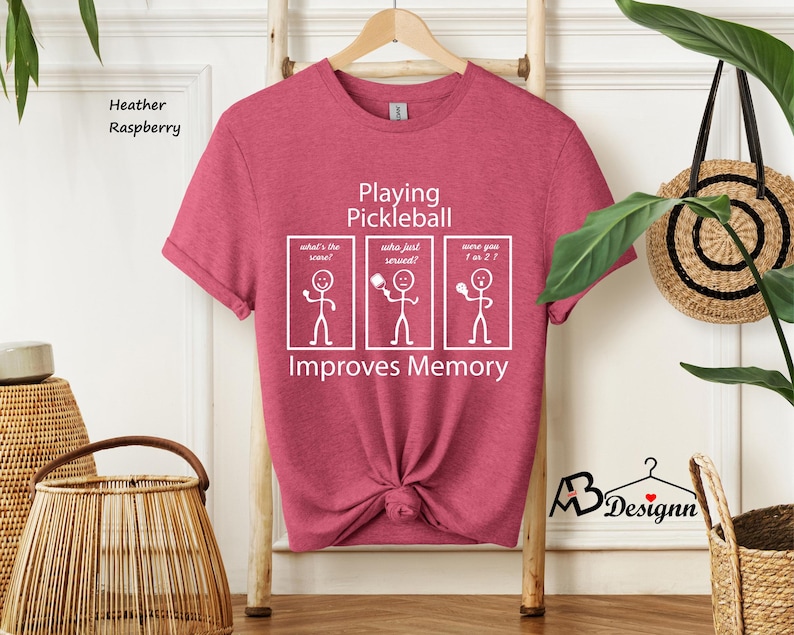 May include: Heather raspberry t-shirt with a white graphic that says "Playing Pickleball Improves Memory". The graphic features stick figures with speech bubbles that say "What's the score?", "Who just served?", and "Were you 1 or 2?"