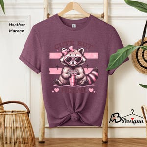 Op de afbeelding: Heather-marron T-shirt met een schattig cartoon-wasbeer die een roze zonnebril draagt en een roze drankje drinkt. De tekst "Cute but psycho" staat op het shirt gedrukt.
