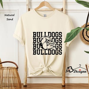 Puede incluir: Una camiseta color crema con la palabra "BULLDOGS" apilada cuatro veces en negro, con un gr&aacute;fico de bulldog. La camiseta se exhibe en un estante de madera. Las palabras "Natural Sand" est&aacute;n impresas en el lado izquierdo.