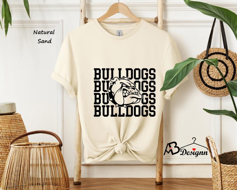 Puede incluir: Una camiseta color crema con la palabra "BULLDOGS" apilada cuatro veces en negro, con un gr&aacute;fico de bulldog. La camiseta se exhibe en un estante de madera. Las palabras "Natural Sand" est&aacute;n impresas en el lado izquierdo.