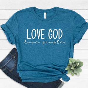 K&ouml;nnte beinhalten: T&uuml;rkisfarbenes T-Shirt mit dem wei&szlig;en Schriftzug "LOVE GOD love people". Das T-Shirt ist am unteren Saum geknotet und mit einer blauen Jeans kombiniert. Das T-Shirt ist aus einem weichen Material.