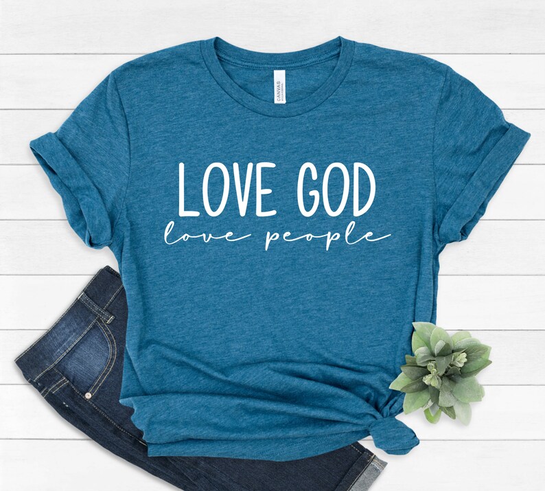 K&ouml;nnte beinhalten: T&uuml;rkisfarbenes T-Shirt mit dem wei&szlig;en Schriftzug "LOVE GOD love people". Das T-Shirt ist am unteren Saum geknotet und mit einer blauen Jeans kombiniert. Das T-Shirt ist aus einem weichen Material.