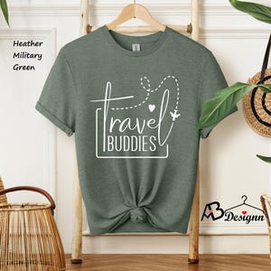 K&ouml;nnte beinhalten: Ein kurz&auml;rmeliges T-Shirt in heather military green mit wei&szlig;em Grafiktext, der travel BUDDIES liest. Das Design enth&auml;lt eine gestrichelte Flugroute, ein kleines Herz und die Silhouette eines Flugzeugs. Das T-Shirt h&auml;ngt an einem Holzkleiderb&uuml;gel.