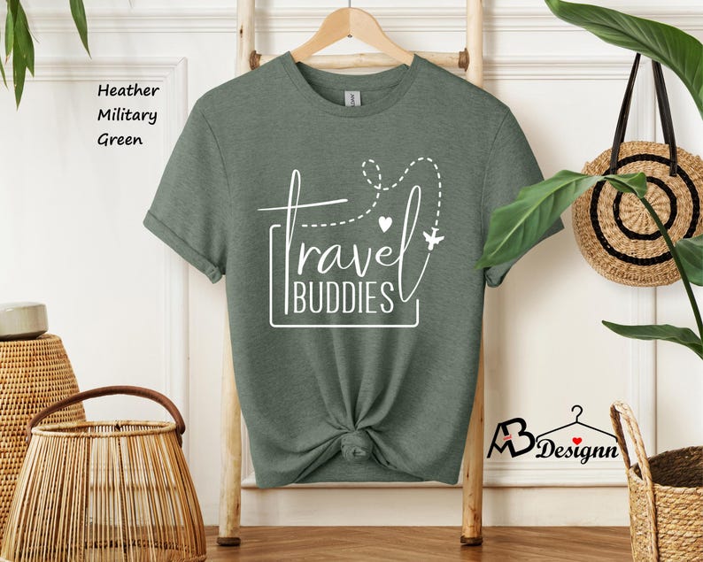 K&ouml;nnte beinhalten: Ein kurz&auml;rmeliges T-Shirt in heather military green mit wei&szlig;em Grafiktext, der travel BUDDIES liest. Das Design enth&auml;lt eine gestrichelte Flugroute, ein kleines Herz und die Silhouette eines Flugzeugs. Das T-Shirt h&auml;ngt an einem Holzkleiderb&uuml;gel.