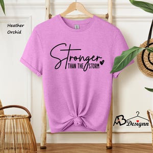 Może przedstawiać: Koszulka w kolorze orchidea z napisem "Stronger than the storm" czarnymi literami z małym sercem.