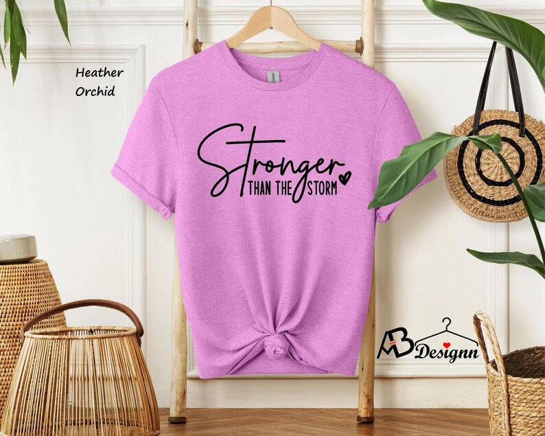 Może przedstawiać: Koszulka w kolorze orchidea z napisem "Stronger than the storm" czarnymi literami z małym sercem.