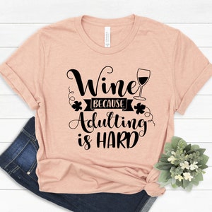 Pu&ograve; includere: Una maglietta color pesca con la scritta "Wine because Adulting is HARD" in corsivo nero. Un grafico di calice di vino &egrave; accanto alla parola "Wine". La maglietta &egrave; piegata con un paio di jeans blu e una piccola pianta.