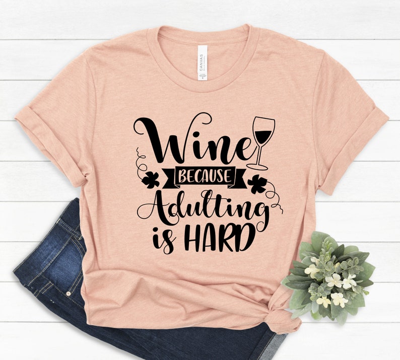 Pu&ograve; includere: Una maglietta color pesca con la scritta "Wine because Adulting is HARD" in corsivo nero. Un grafico di calice di vino &egrave; accanto alla parola "Wine". La maglietta &egrave; piegata con un paio di jeans blu e una piccola pianta.