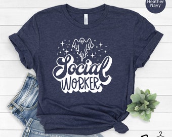 May include: Heather navy blue t-shirt with the words "Social Worker" in a white, stylized font, accompanied by a ghost graphic and stars. The shirt is folded and styled with a knot at the bottom, and is paired with blue jeans.