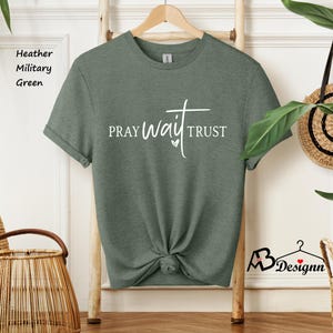 May include: Heather military green t-shirt with the words "PRAY WAIT TRUST" in white script. The shirt is knotted at the waist and hangs on a wooden hanger. The shirt is displayed in a well-lit room.