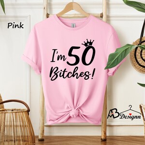 May include: A light pink short-sleeved t-shirt featuring black text that reads I'm 50 Bitches! with a small crown graphic above the number 50. The shirt has a crew neckline and is displayed on a wooden hanger, with the bottom hem tied in a knot.