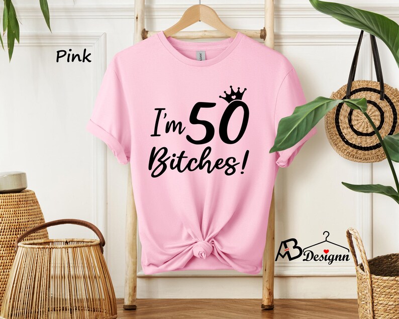 May include: A light pink short-sleeved t-shirt featuring black text that reads I'm 50 Bitches! with a small crown graphic above the number 50. The shirt has a crew neckline and is displayed on a wooden hanger, with the bottom hem tied in a knot.