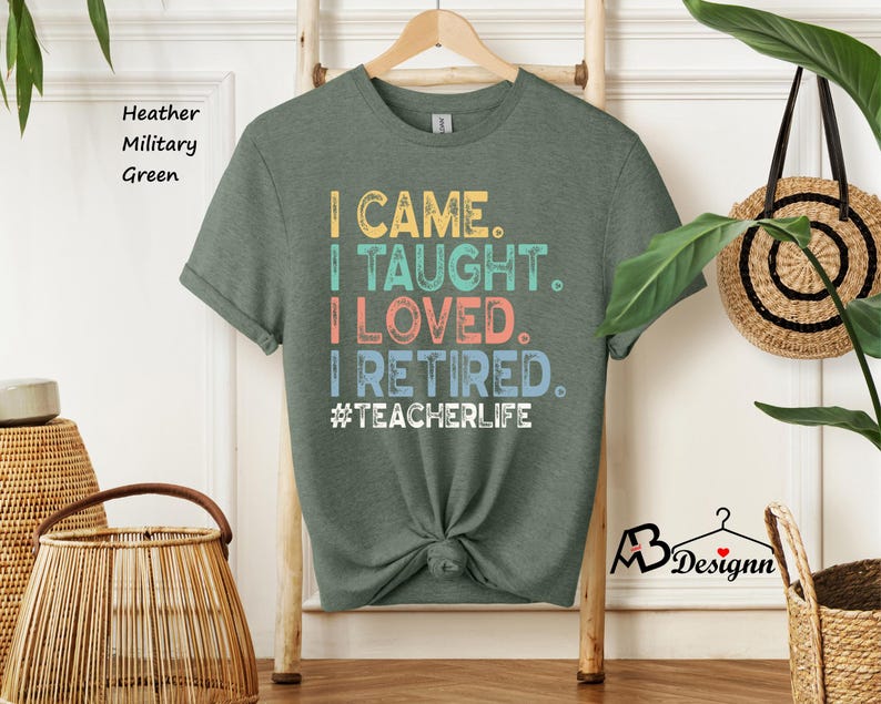 Może przedstawiać: Heather wojskowa zielona koszulka z napisem "I CAME. I TAUGHT. I LOVED. I RETIRED. #TEACHERLIFE" w kolorowych literach. Koszulka jest zawiązana w pasie i wisi na drewnianym wieszaku.