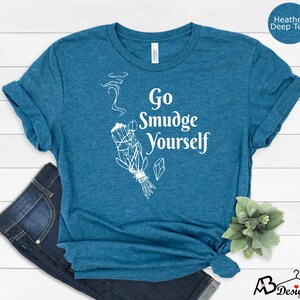 K&ouml;nnte beinhalten: Ein blaues T-Shirt mit dem Text "Go Smudge Yourself" in Wei&szlig;. Das Shirt zeigt eine wei&szlig;e Illustration eines R&auml;ucherstabs mit aufsteigendem Rauch.