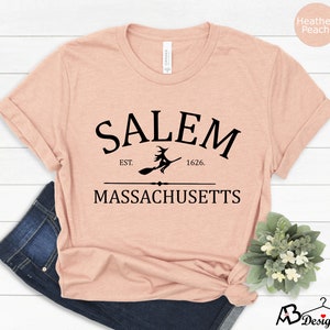 May include: A heather peach t-shirt with a black graphic design that says "Salem Est. 1626. Massachusetts" with a silhouette of a witch flying on a broomstick.