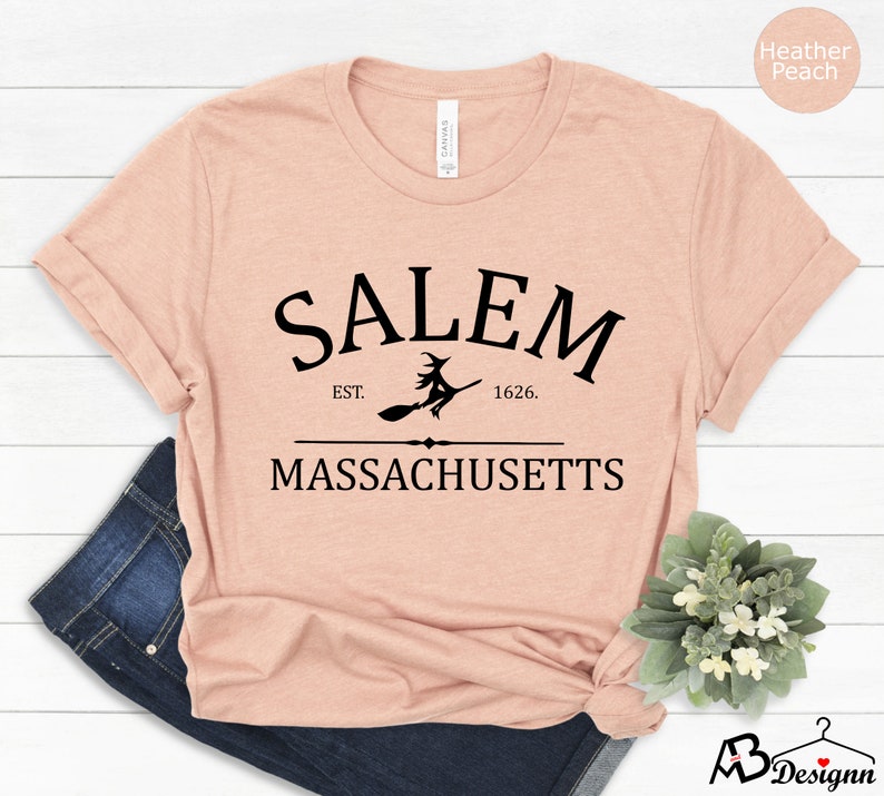 May include: A heather peach t-shirt with a black graphic design that says "Salem Est. 1626. Massachusetts" with a silhouette of a witch flying on a broomstick.