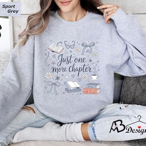 May include: Light grey sweatshirt with the text "Just one more chapter" and illustrations of books, glasses, bows, and flowers. The sweatshirt is a sport grey color.