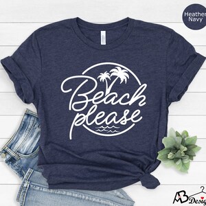 Może przedstawiać: Koszulka w kolorze granatowym melanżowym z białym kołem z napisem "Beach Please" i palmami oraz falami wewnątrz koła.