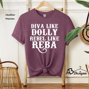 Puede incluir: Una camiseta de manga corta en color granate jaspeado con el texto en blanco que dice DIVA LIKE DOLLY REBEL LIKE REBA. La prenda tiene cuello redondo y est&aacute; colgada en una percha de madera. La tela tiene una apariencia texturizada y jaspeada.
