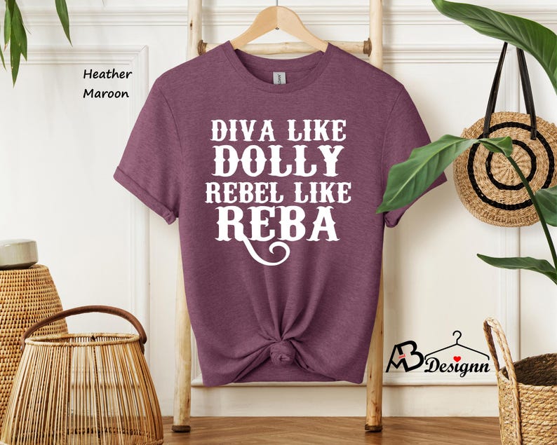 Puede incluir: Una camiseta de manga corta en color granate jaspeado con el texto en blanco que dice DIVA LIKE DOLLY REBEL LIKE REBA. La prenda tiene cuello redondo y est&aacute; colgada en una percha de madera. La tela tiene una apariencia texturizada y jaspeada.