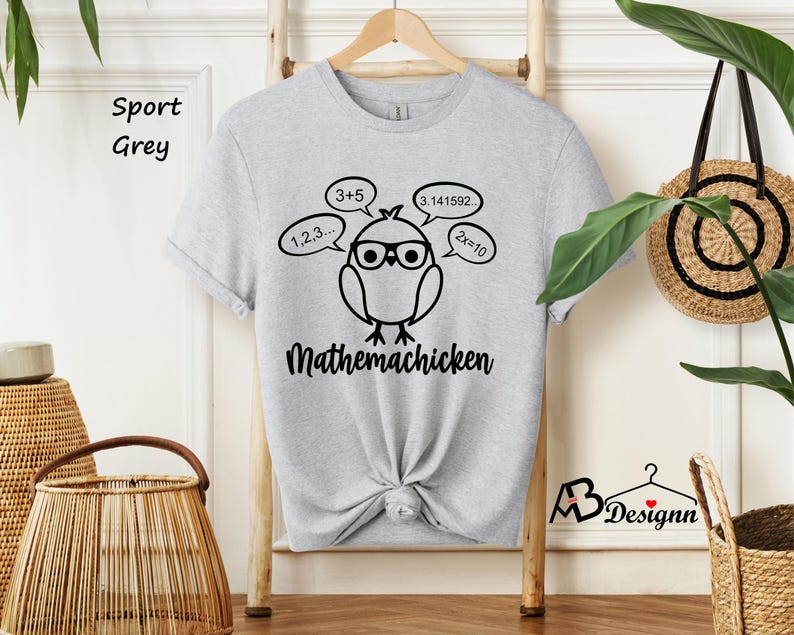 Op de afbeelding: Een grijs T-shirt met een zwarte afbeelding van een cartoonkip met een bril en omringd door wiskundige vergelijkingen. De tekst "Mathemachicken" staat onder de kip.
