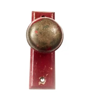 May include: A red door knob with a brass finish. The knob is round and has a small hole in the center. The door knob is attached to a red metal plate.