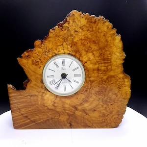 May include: A wooden clock with a natural burl finish. The clock face is white with black Roman numeral hour markers and black hands. The clock is made by the company "Dugena".