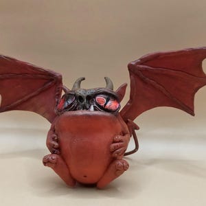 May include: A red, cartoonish gargoyle figurine with large red wings, horns, and glowing red eyes. The figurine is in a seated position, with its hands clasped in front. The overall design is whimsical and detailed.