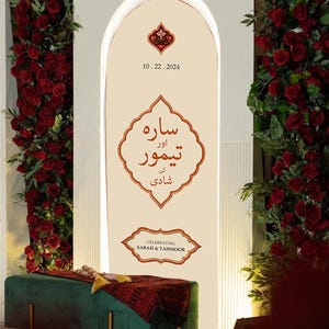 Op de afbeelding: Een witte boog met een crèmekleurige achtergrond en gouden afwerking. De boog is versierd met rode rozen en een bord met de tekst "Sara & Taimoor" en "Viering 10.22.2024".