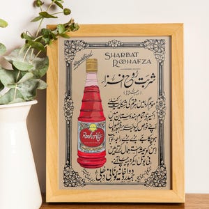 May include: A vintage advertisement for Rooh Afza Sharbat, a rose-flavored syrup. The ad features a bottle of the syrup with the brand name and text in Urdu. The ad is framed in a light wood frame.