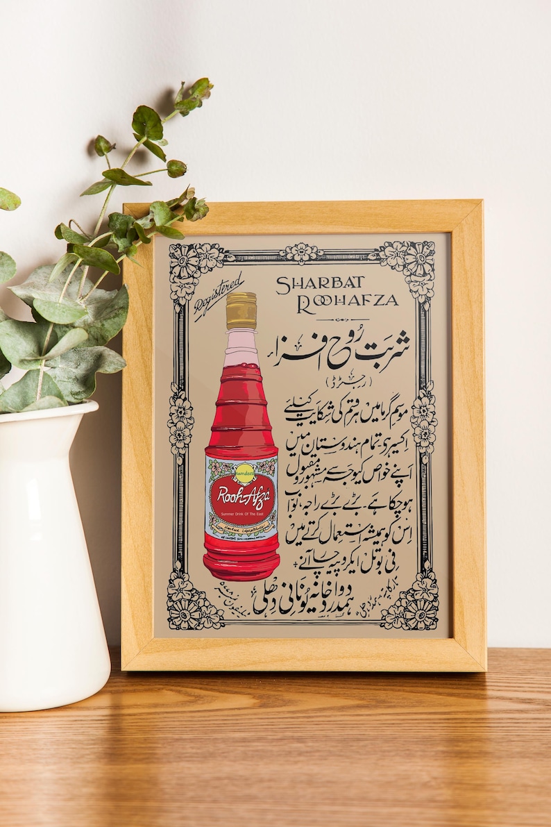 May include: A vintage advertisement for Rooh Afza Sharbat, a rose-flavored syrup. The ad features a bottle of the syrup with the brand name and text in Urdu. The ad is framed in a light wood frame.