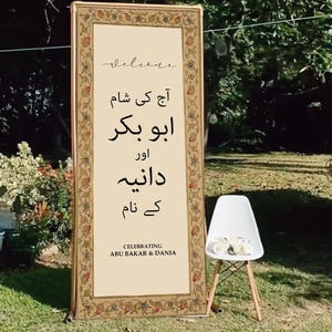 Op de afbeelding: Decoratief welkomstbord met een gouden lijst en bloemenrand. Het bord bevat tekst in een vreemde taal en de Engelse tekst "CELEBRATING ABU BAKAR & DANIA". Een witte stoel met bloemen staat in de buurt.