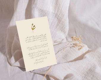 May include: A cream-colored card with calligraphy and a gold floral design, resting on a white, textured fabric. The card features text in a script, and a dried flower stem is placed near it.