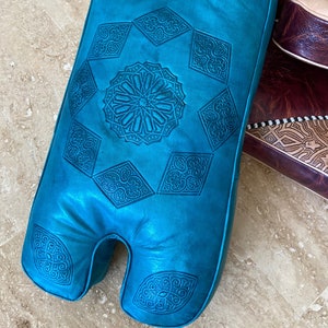 May include: A teal blue leather pouf with intricate embossed patterns. The pouf has a unique shape with two separate sections for sitting.