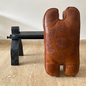 May include: A brown leather pouf with intricate embossed designs. The pouf has a unique shape with two rounded ends and a center opening. The pouf is sitting on a wooden floor.