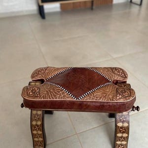 May include: A brown leather Moroccan pouf with intricate floral carvings. The pouf has a square top and four legs made of wood with a light brown finish.