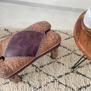 Peut inclure: Un tabouret en cuir marron fait à la main, orné de motifs en relief complexes. Le centre présente un panneau en cuir bordeaux foncé avec un motif floral. Le tabouret a une forme unique et est posé sur un tapis à motifs.