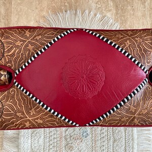 May include: A red leather pouf with a diamond-shaped top and intricate brown leather designs. The pouf has a black and white trim around the edges.