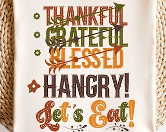 May include: Cream-colored t-shirt with the words "THANKFUL, GRATEFUL, BLESSED, HANGRY! Let's Eat!" in a fall-themed color palette. The shirt is folded on a woven, circular placemat.