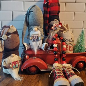 Puede incluir: Tres gnomos con fotos personalizadas, uno sentado en un cami&oacute;n de juguete rojo con un saco de arpillera que dice "Comida para renos" y una guirnalda de cuentas negras y rojas. Los gnomos llevan sombreros y tienen barbas blancas.
