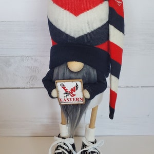 Puede incluir: Un gnomo con un gorro a rayas rojo, blanco y azul y un su&eacute;ter negro sostiene un cartel que dice "Eastern Washington University". El gnomo lleva zapatillas negras con cordones blancos.