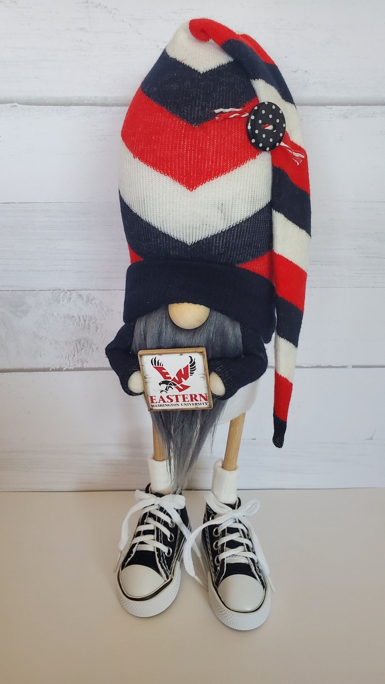 Puede incluir: Un gnomo con un gorro a rayas rojo, blanco y azul y un su&eacute;ter negro sostiene un cartel que dice "Eastern Washington University". El gnomo lleva zapatillas negras con cordones blancos.