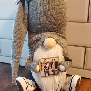 Puede incluir: Un gnomo gris con barba blanca sosteniendo una foto enmarcada de una familia de cuatro. El gnomo lleva un gorro gris y zapatos blancos.