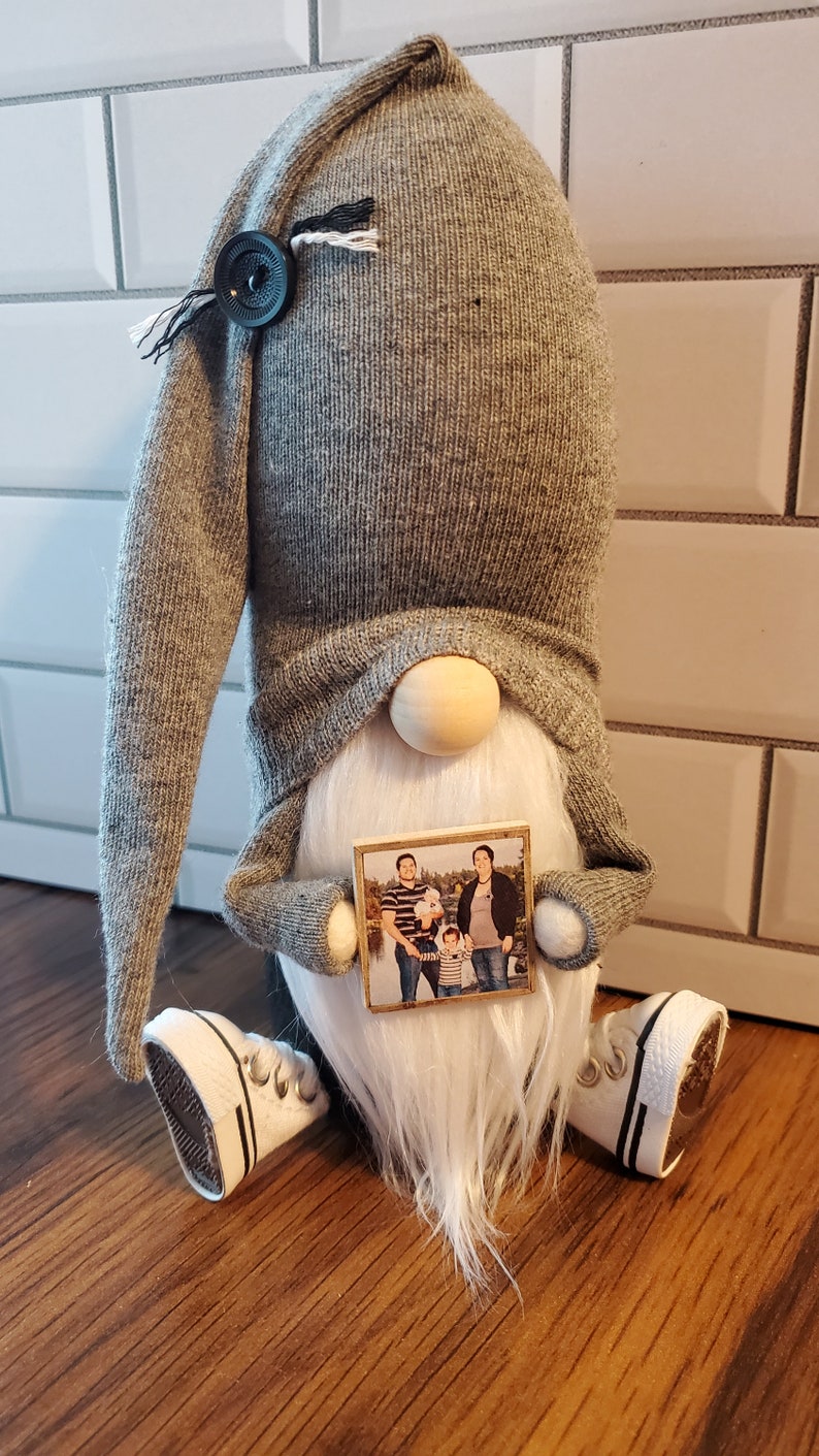 Puede incluir: Un gnomo gris con barba blanca sosteniendo una foto enmarcada de una familia de cuatro. El gnomo lleva un gorro gris y zapatos blancos.