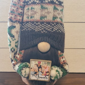 Puede incluir: Un gnomo con un gorro de punto marr&oacute;n con un bot&oacute;n blanco y un dise&ntilde;o estampado verde y marr&oacute;n. El gnomo sostiene un peque&ntilde;o letrero de madera con una foto de una familia y el texto "The Grands".