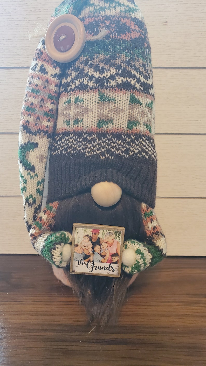 Puede incluir: Un gnomo con un gorro de punto marr&oacute;n con un bot&oacute;n blanco y un dise&ntilde;o estampado verde y marr&oacute;n. El gnomo sostiene un peque&ntilde;o letrero de madera con una foto de una familia y el texto "The Grands".