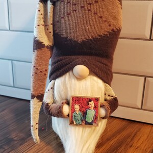 Puede incluir: Un gnomo de rayas marrones y blancas con barba blanca sostiene una foto enmarcada de dos ni&ntilde;os frente a un arbusto de flores rojas. El gnomo lleva un gorro de rayas marrones y blancas con un bot&oacute;n en la parte superior.