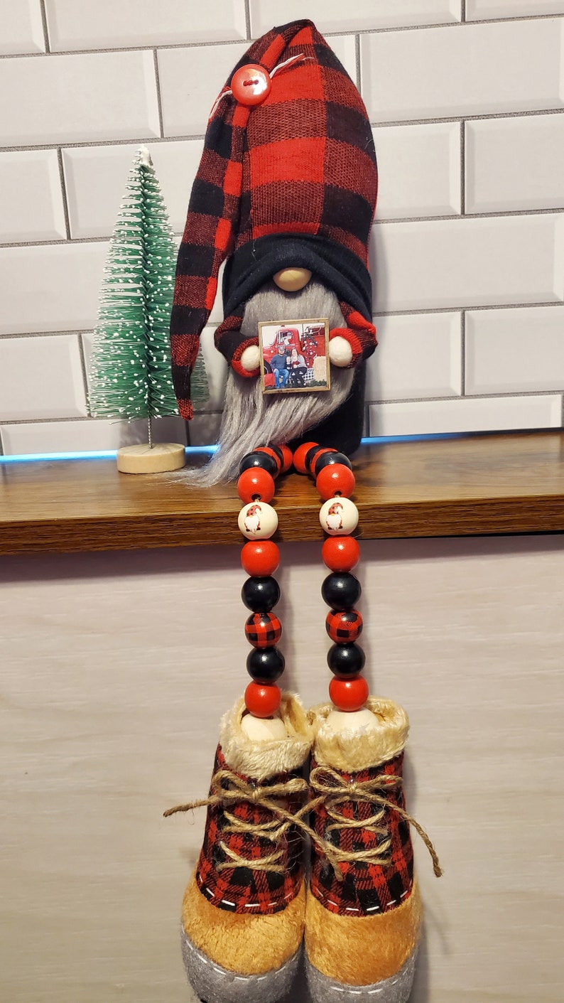 Puede incluir: Un gnomo de cuadros rojos y negros con una larga barba blanca est&aacute; sentado con piernas hechas de cuentas de madera. El gnomo lleva un gorro de cuadros rojos y negros y sostiene una foto de una familia frente a un cami&oacute;n rojo. El gnomo lleva botas marrones con detalles de cuadros rojos y negros.