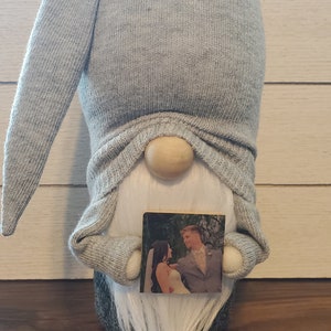 Puede incluir: Un gnomo gris con barba blanca sosteniendo una peque&ntilde;a foto de una novia y un novio. El gnomo lleva un gorro gris con un bot&oacute;n negro.